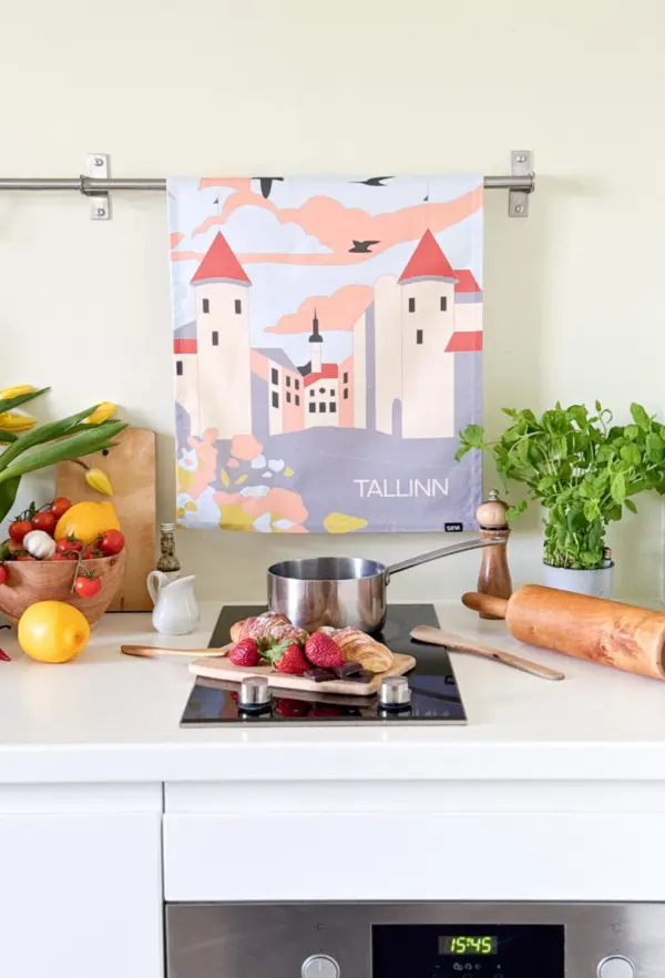 A modern kitchen worktop features a saucepan, strawberries on a chopping board, and a New Rebels small rucksack. Behind them hangs the "Köögirätik – Viru väravad" tea towel, with produce in a paper bag, potted herb, pepper mill, and rolling pin nearby. Gate21.ee