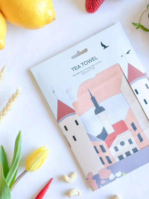A packaged Köögirätik – Viru väravad tea towel displays buildings with red roofs, a church steeple, lemons, a strawberry, a yellow tulip, green leaves, cashew nuts, and a stylish New Rebels small rucksack on a white surface. Gate21.ee
