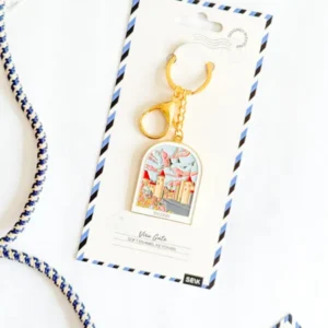 A gold keyring featuring a colourful illustrated charm from the Võtmehoidja – Talvine mets collection is displayed on a white card with blue striped edges. The card, labelled “Vienna, Austria” and “Soft enamel keyring,” evokes the modern design often seen in New Rebels small rucksack collections. The item is shown resting on a white surface. Gate21.ee