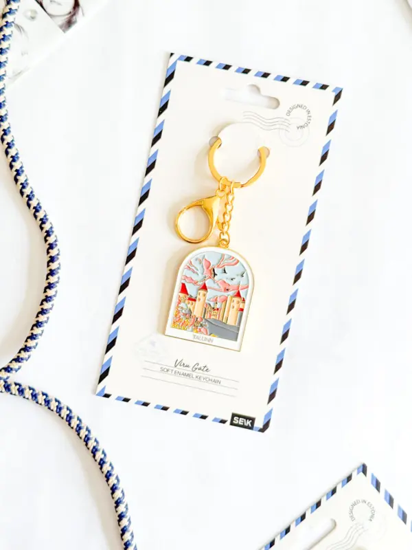 A gold keyring featuring a colourful illustrated charm from the Võtmehoidja – Talvine mets collection is displayed on a white card with blue striped edges. The card, labelled “Vienna, Austria” and “Soft enamel keyring,” evokes the modern design often seen in New Rebels small rucksack collections. The item is shown resting on a white surface. Gate21.ee