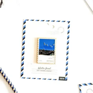 A rectangular enamel Magnet – talvine mets, featuring an illustration of a winter forest with birds, is attached to a postcard-style backing labelled Estonia. Beside the card, a blue and white cord rests on a white surface, evoking the cosy charm you might feel while packing your New Rebels small rucksack for a crisp northern adventure. Gate21.ee