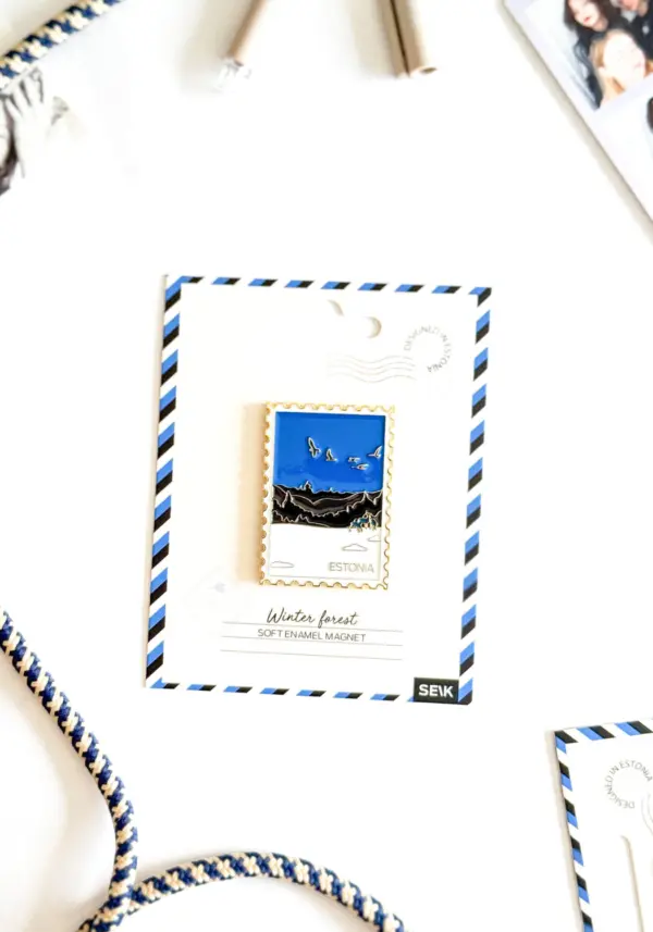 A rectangular enamel Magnet – talvine mets, featuring an illustration of a winter forest with birds, is attached to a postcard-style backing labelled Estonia. Beside the card, a blue and white cord rests on a white surface, evoking the cosy charm you might feel while packing your New Rebels small rucksack for a crisp northern adventure. Gate21.ee
