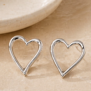 A pair of Love - BØLD Earrings - Silver is displayed on a beige stone surface, with part of a round ceramic dish visible in the background. Gate21.ee