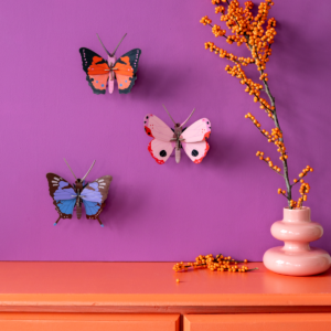 Three decorative Rosora Butterflies from StudioROOF are mounted on a purple wall above an orange chest of drawers. A white vase with orange berries sits on the chest of drawers, with some berries scattered beside it. Gate21.ee