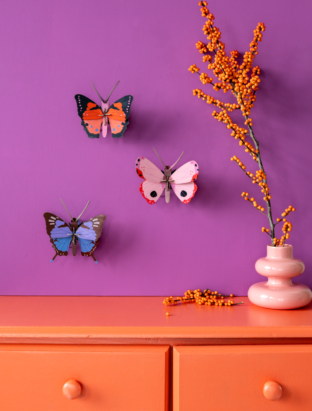 Three decorative Rosora Butterflies from StudioROOF are mounted on a purple wall above an orange chest of drawers. A white vase with orange berries sits on the chest of drawers, with some berries scattered beside it. Gate21.ee
