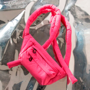 A Knotflex Pack - Magenta, featuring two thick straps, rests on a reflective crumpled metallic surface. The bright pink bag has a visible zip pocket and boasts a soft, plush design that stands out for its unique style. Gate21.ee