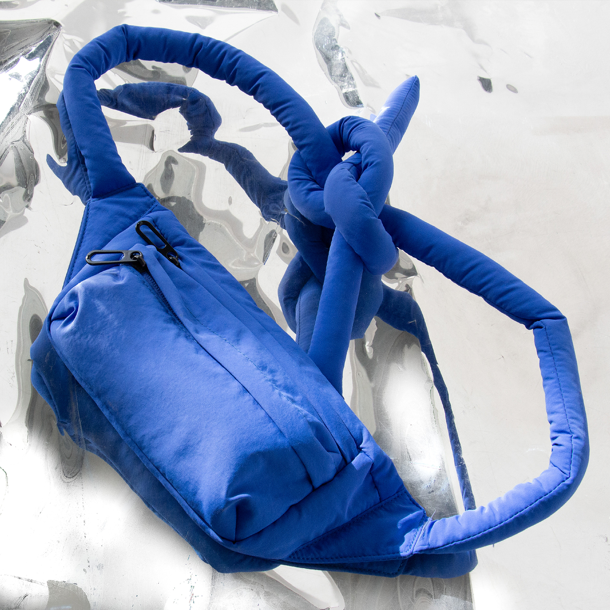 A magenta Knotflex Pack with a padded design and a front zip pocket is placed on a shiny, reflective silver surface. The thick strap is tied in a loose knot. Gate21.ee