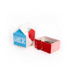 A ceramic &ldquo;Milk - Jewellery Box&rdquo; shaped like a strawberry milk carton sits with its lid off, revealing small jewellery items inside. Reminiscent of the whimsical Dreamy cat jewellery plate by Hello Ferretti, the lid displays a strawberry image and the words &ldquo;Strawberry Milk.&rdquo; A beaded bracelet rests on the edge of the box. Gate21.ee