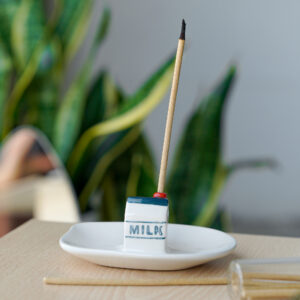 A ceramic incense burner, named “Incense burner - Milk,” is shaped like a small milk carton and sits atop a white dish. Reminiscent of Helio Ferretti’s modern and colourful designs, an incense stick is placed inside the holder, with a plant and softly blurred objects in the background on a light wooden surface. Gate21.ee