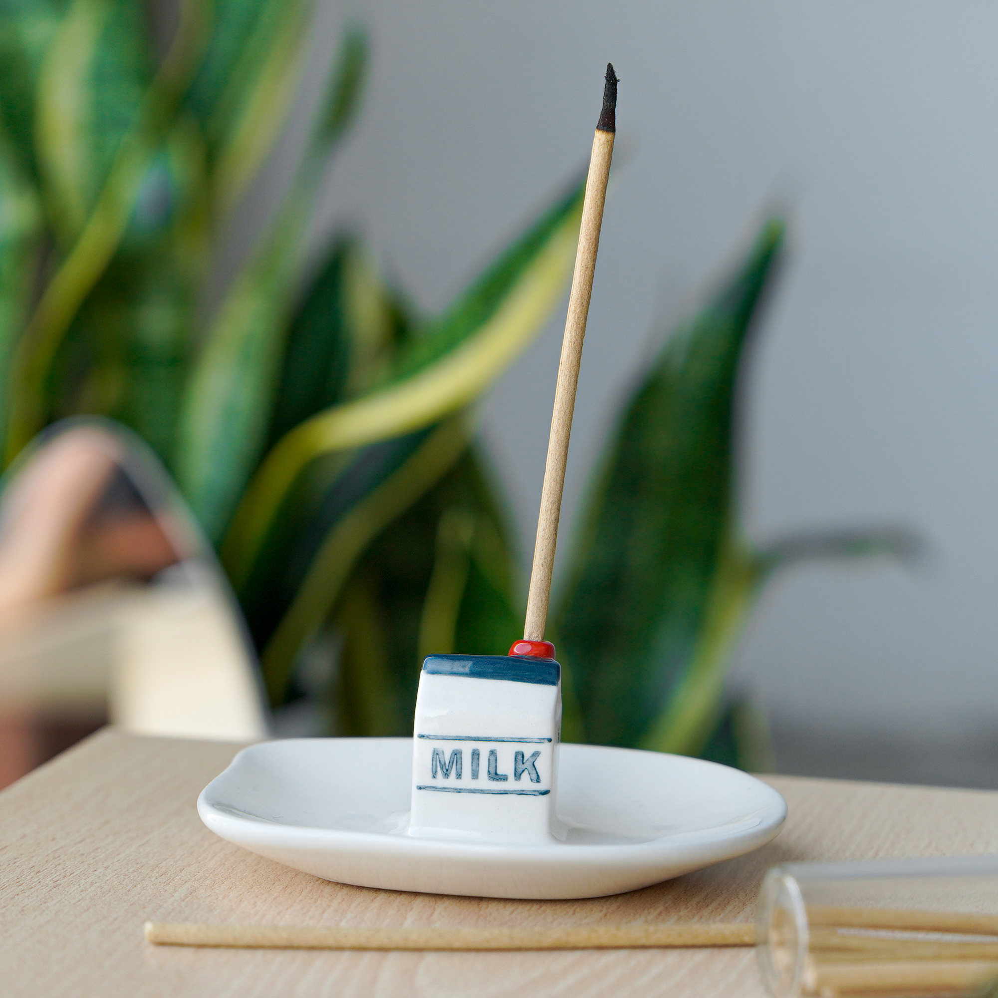 A ceramic incense burner, named “Incense burner - Milk,” is shaped like a small milk carton and sits atop a white dish. Reminiscent of Helio Ferretti’s modern and colourful designs, an incense stick is placed inside the holder, with a plant and softly blurred objects in the background on a light wooden surface. Gate21.ee