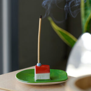 A burning incense stick stands upright in the Incense burner - Milk, a small ceramic holder shaped like a house on a green base, with smoke rising. A plant and part of a metallic object are visible in the background, highlighting the playful charm of modern and colourful incense burners by Helio Ferreti. Gate21.ee