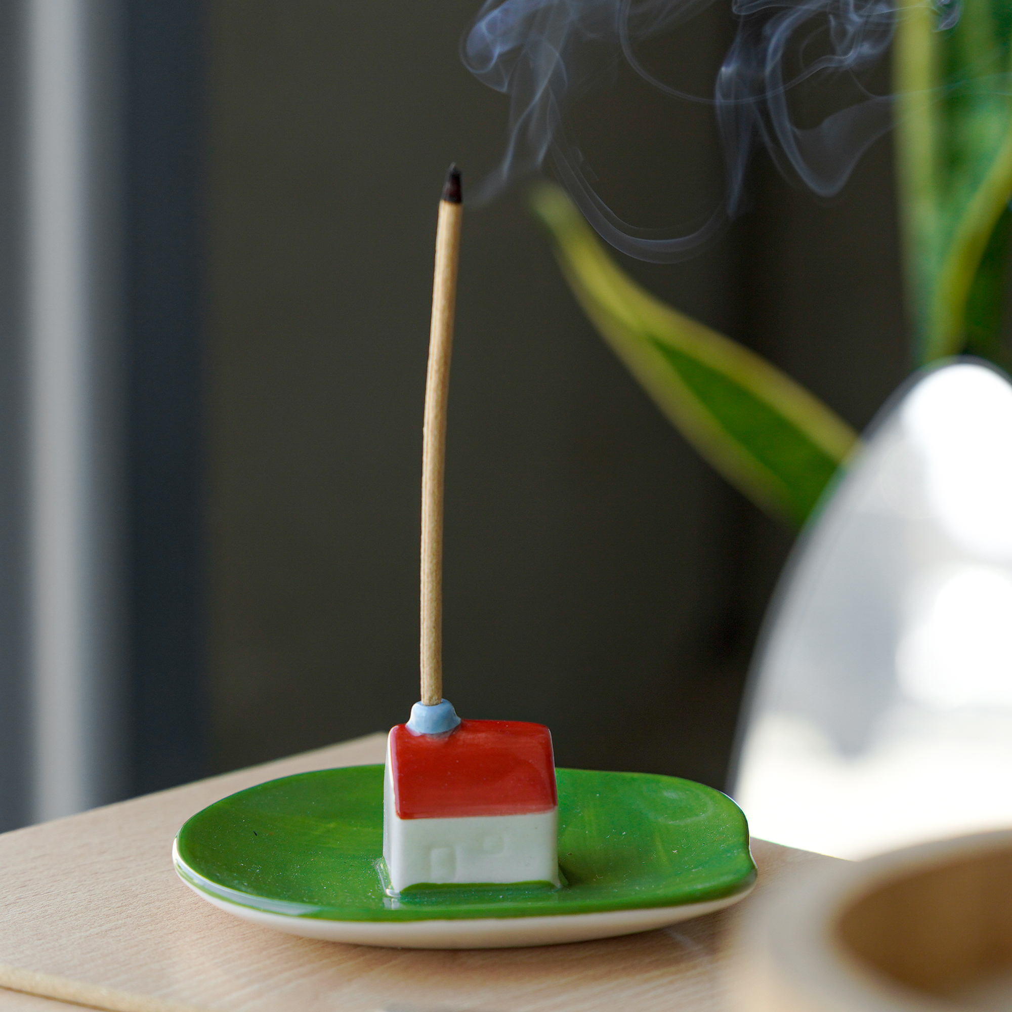 A burning incense stick stands upright in the Incense burner - Milk, a small ceramic holder shaped like a house on a green base, with smoke rising. A plant and part of a metallic object are visible in the background, highlighting the playful charm of modern and colourful incense burners by Helio Ferreti. Gate21.ee
