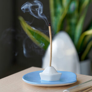 A lit incense stick with smoke gently rising from its tip sits in a white, boat-shaped holder on a blue dish, placed atop a wooden surface. In the background, lush green foliage adds to the tranquil ambience, perfectly complementing the modern and colourful Incense burner - Milk by Helio Ferreti. Gate21.ee