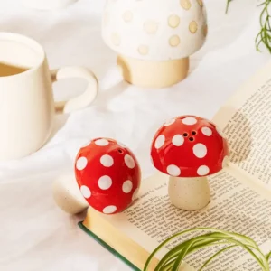 Mushroom - Salt & Pepper Shakers in red and white rest on an open book, joined by a ceramic cup, a white mushroom decor piece, and green plant leaves atop a white surface—an enchanting kitchen accent perfect for any space. Gate21.ee