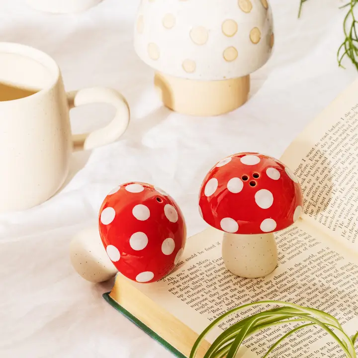 Mushroom - Salt & Pepper Shakers in red and white rest on an open book, joined by a ceramic cup, a white mushroom decor piece, and green plant leaves atop a white surface—an enchanting kitchen accent perfect for any space. Gate21.ee
