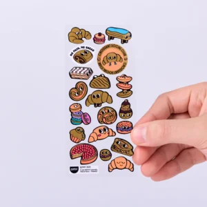 A hand holds a sheet of Criossant - Fabulous Stickers, featuring colourful cartoon bread and pastry characters like croissants, rolls, and doughnuts. The top sticker reads “NO PAIN, NO BREAD” and “CROISSANT SOCIAL CLUB.” Perfect for decorating your OMY sticker earrings or personalising notebooks and gadgets with adorable bakery vibes. Gate21.ee