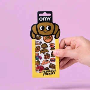 A hand holds a yellow packet labelled “Criossant - Fabulous Stickers,” featuring assorted illustrated pastry and croissant stickers. The packaging displays a cartoon croissant character at the top against a pink background, giving it a playful look similar to OMY sticker earring designs. Gate21.ee