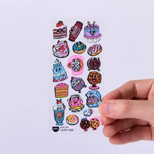 A hand holds a sheet of Donut - Fabulous Stickers, featuring colourful cartoon desserts like cakes, doughnuts, cupcakes, and milkshakes. Some stickers include fun phrases such as DOWN TO BE SWEET. These playful stickers add an extra touch of fun to any collection. Gate21.ee