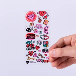 A hand holds a sheet of the Heart - Fabulous Stickers, showcasing colourful designs like hearts, cherries, lips, a key, hands, cheerful cartoon characters, and playful text such as LOVE, HUGS, and MON AMOUR—perfect for creating your own OMY sticker earrings or adding a fun touch to your belongings. Gate21.ee