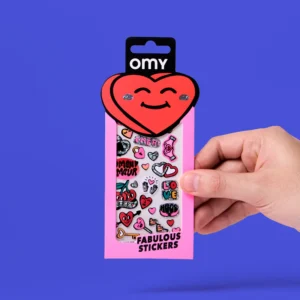 A hand holds a packet of Heart - Fabulous Stickers. The packaging is pink with a smiling heart-shaped top, featuring playful designs and colourful hearts. The stickers include fun words and patterns reminiscent of OMY sticker earrings, all set against a vibrant blue background. Gate21.ee