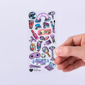 A hand holds a sheet of Lashes - Fabulous Stickers, featuring colourful designs like OMY sticker earrings, make-up items, eyes, hearts, and words such as “self care” and “slay,” all set against a light purple background. Gate21.ee
