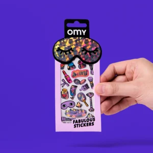 A hand holds a pink packet labelled “Lashes - Fabulous Stickers,” showcasing make-up-themed stickers and a pair of OMY sticker earrings, with a shiny, multicoloured sleep mask attached at the top. The background is solid purple. Gate21.ee