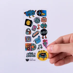 A hand holds a sheet of Music – Fabulous Stickers displaying vibrant images of musical items like ghetto blasters, records, headphones, and musical notes, along with cartoon faces and playful OMY sticker earrings, all under the title Vibe Check. Gate21.ee