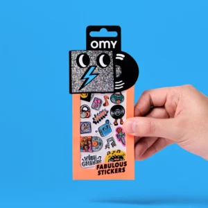 A hand holds a packet of Music - Fabulous Stickers featuring music-themed and retro cartoon designs, reminiscent of playful OMY sticker earrings. The orange packaging showcases a shiny black square character and a vinyl record graphic at the top, set against a blue background. Gate21.ee