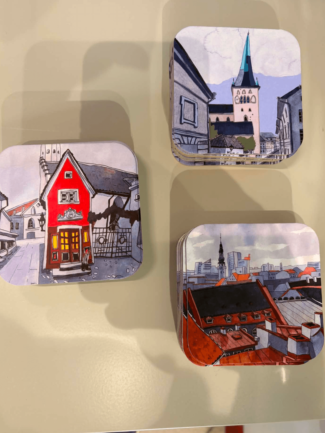Old Town Coasters - Olevuse Art - KÕIK: Set of three square coasters featuring charming illustrated cityscapes—a red house, a church with a tall spire, and rooftops against a skyline—arranged on a light-coloured surface. Gate21.ee
