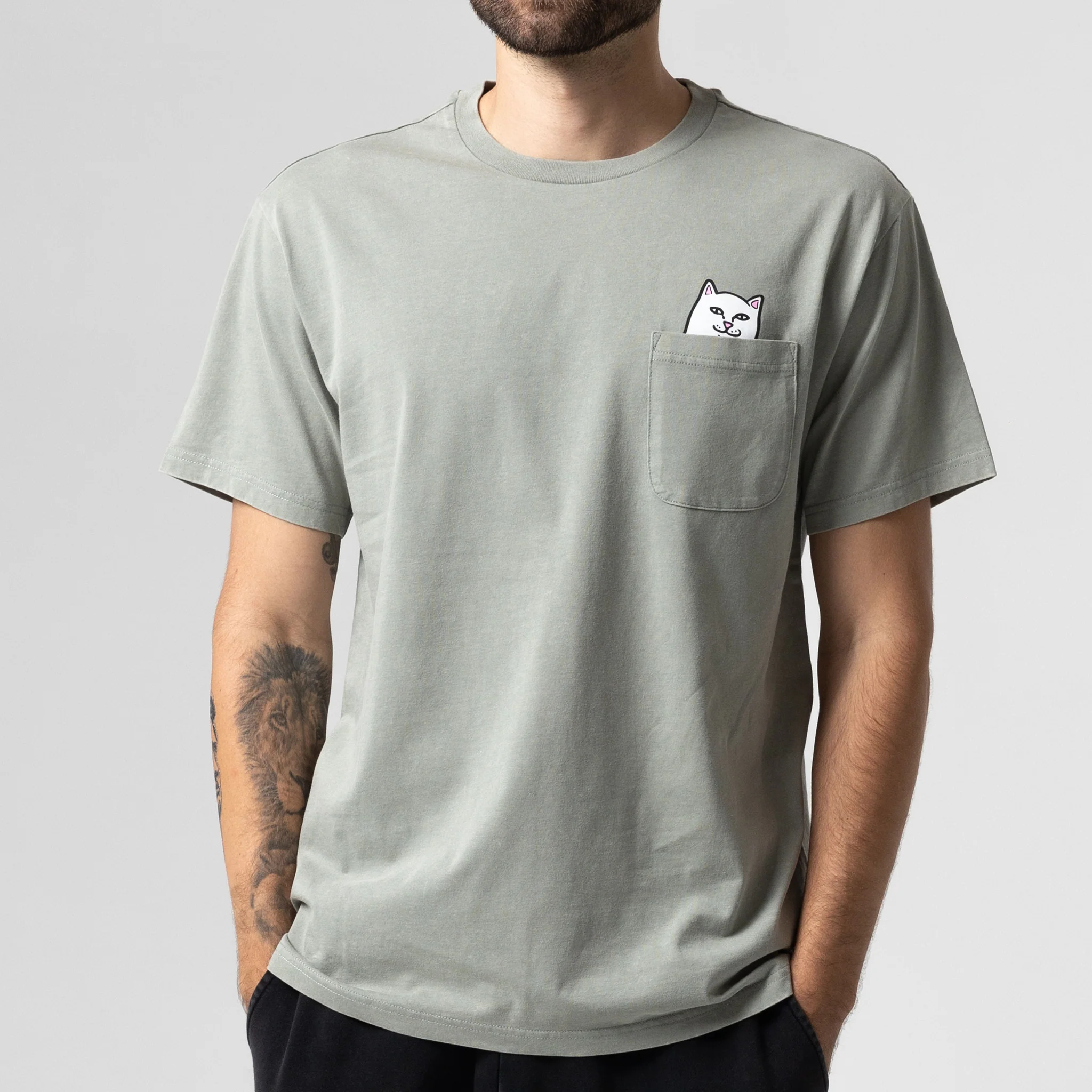 A person wearing a sage vintage wash Lord Nermal Pocket Tee with a small chest pocket featuring an embroidered cartoon cat peeking out, standing against a plain background. Gate21.ee