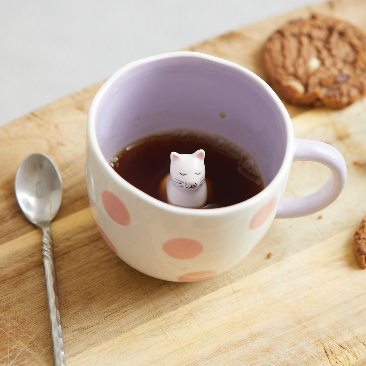 A Surprise Cat - Mug with a pink polka dot design and lavender interior holds tea or coffee, featuring a small ceramic cat figurine sitting inside the mug. A spoon and two biscuits rest on a wooden surface beside the mug. Gate21.ee