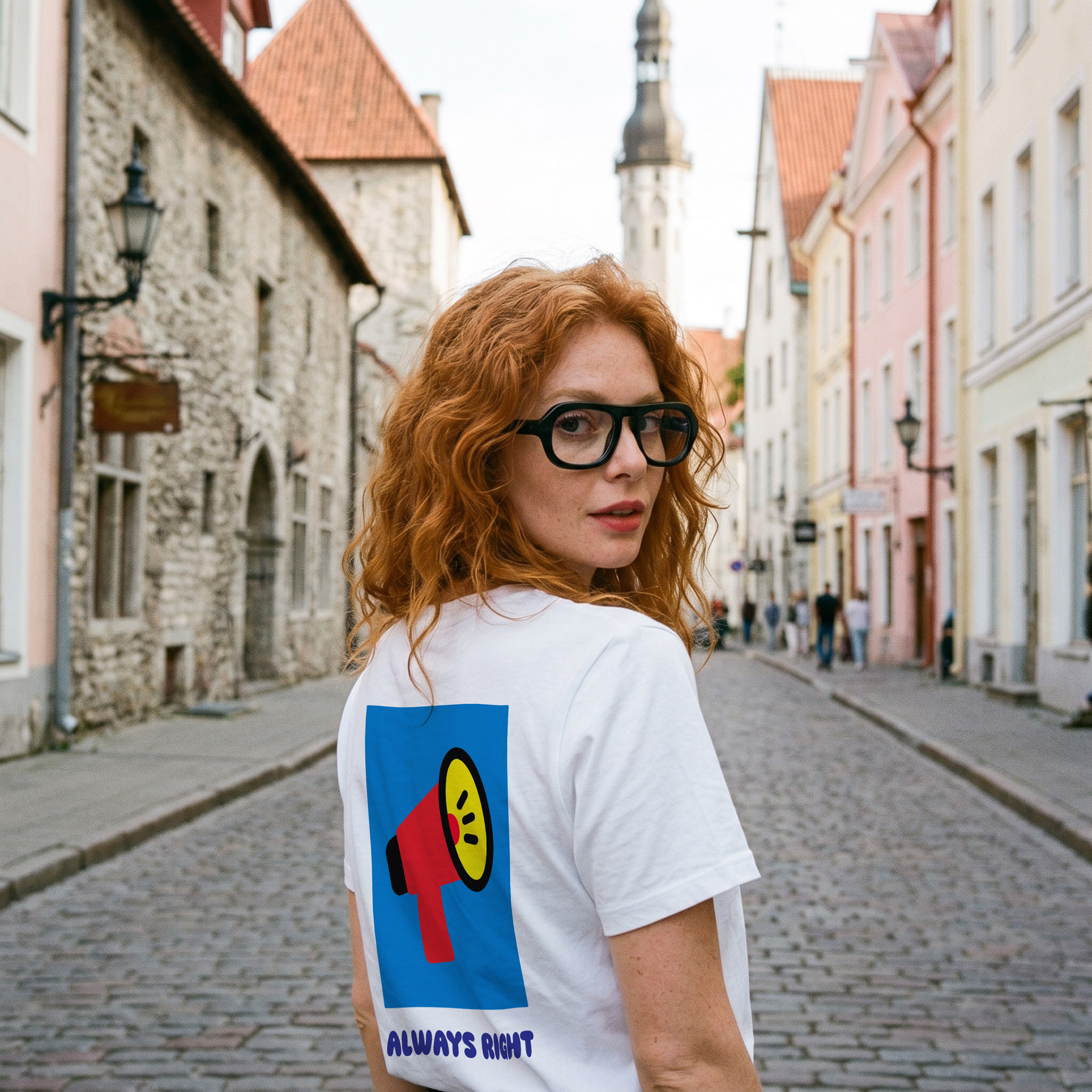 A woman with red hair and glasses stands on a cobbled street lined with pastel buildings. She is wearing the “Always Right - PROJECTION Ts,” a white T-shirt featuring a bold graphic and the text ALWAYS RIGHT on the back, as she looks over her shoulder. Gate21.ee