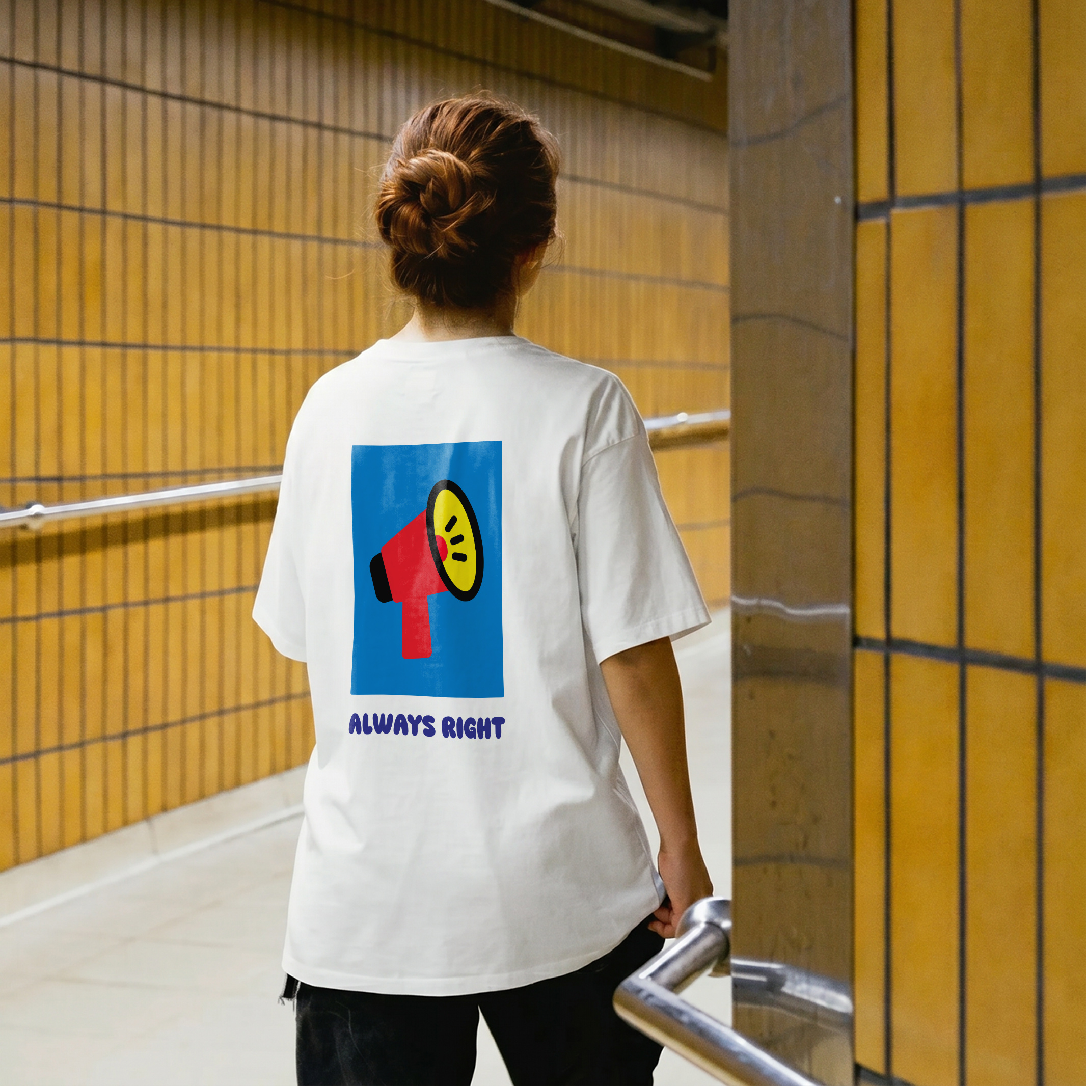 Wearing the Always Right - PROJECTION Ts, which features a megaphone graphic and the text ALWAYS RIGHT on the back, a person with reddish hair in a bun stands in a corridor with yellow tiled walls and metal railings. Gate21.ee