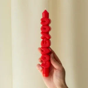 A hand holds the Love You 3D Candle - Red, shaped to spell out “LOVE YOU” vertically. The background is a light beige curtain. Gate21.ee