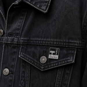 Close-up of a black denim jacket featuring metal buttons and a Good Sign - Pin enamel badge on the pocket flap, which reads WELL THAT’S A GOOD SIGN with a simple stick figure illustration. Gate21.ee