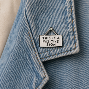 A close-up of a light blue denim jacket with a "This is fine" enamel pin and the Positive Sign - Pin, which features "THIS IS A POSITIVE SIGN" in black letters on a white, hanging-sign style background. Gate21.ee