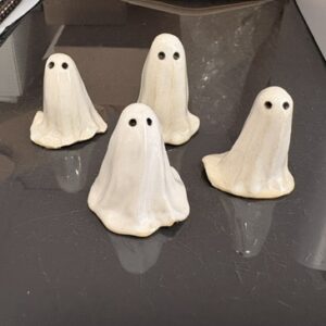 Four large, white ceramic &ldquo;Old Town ghosts&rdquo; figurines from Lily Strider, each with black eyes, are arranged on a shiny black surface that reflects their spectral forms. In the background, scattered papers and a white box contribute an air of mysterious intrigue reminiscent of Lily Strider&rsquo;s tales. Gate21.ee