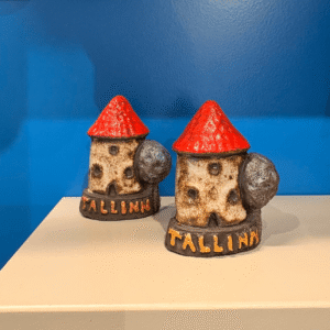 Two Raivo Koit ceramics - Tower Tallinn ceramic souvenirs with red roofs and "Tallinn" written on them are displayed on a shelf against a blue background. Gate21.ee