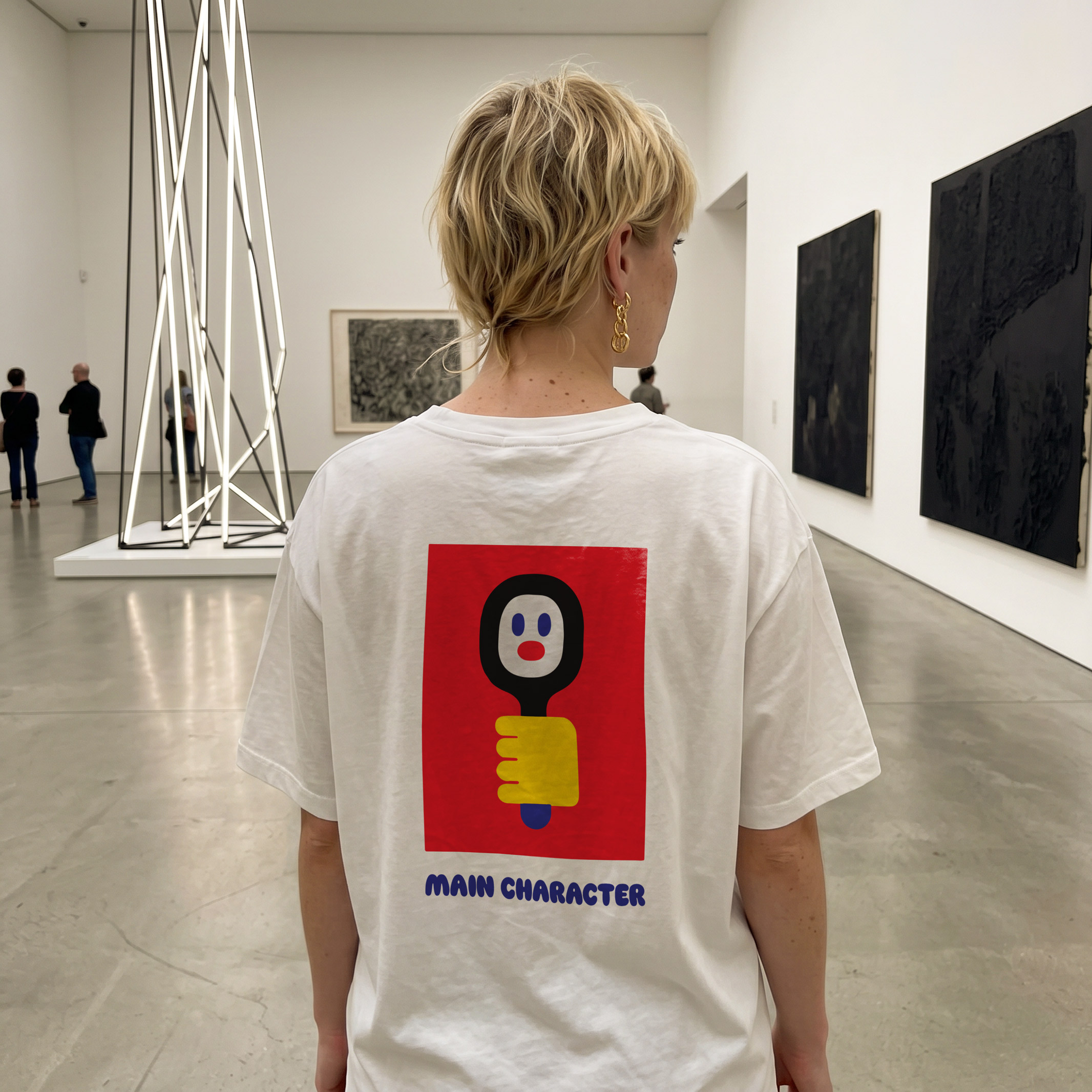 A person with short blond hair is wearing the Main Character - PROJECTION T-shirt, which features a cartoon graphic and the phrase “MAIN CHARACTER,” whilst standing in a modern art gallery adorned with abstract artwork. Gate21.ee