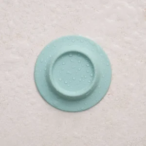 A Soapi - Mint, featuring a light blue circular design with a raised ring and centre and covered in water droplets, is placed on a textured, off-white surface. Gate21.ee