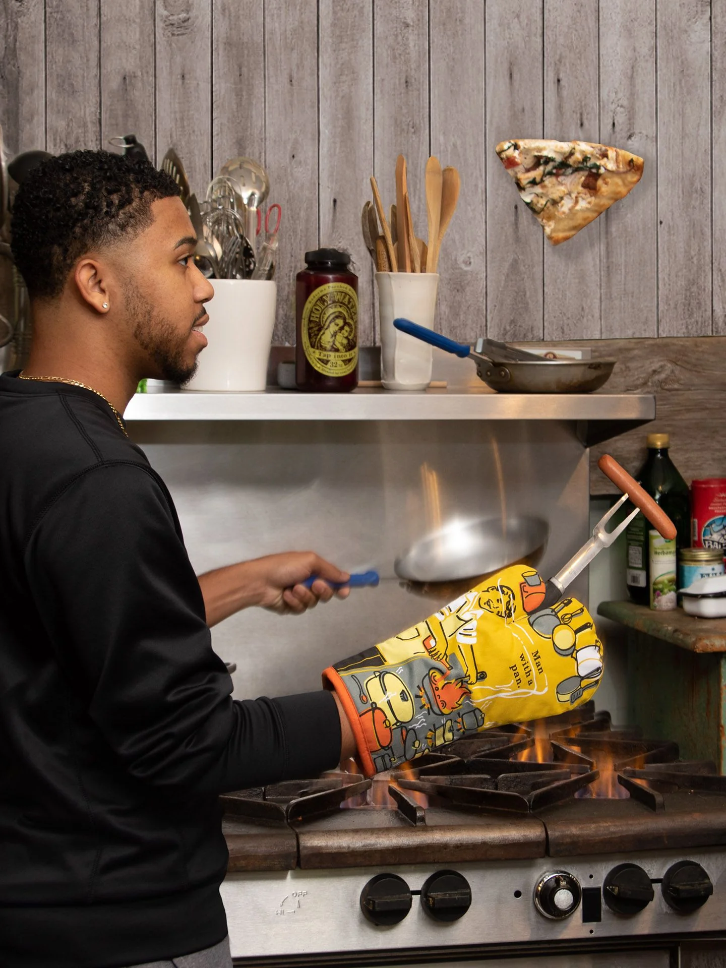A man wearing the “Man with a pan - Oven Glove” flips a frying pan over the hob, sending a slice of pizza airborne, while kitchen utensils and jars decorate the worktop and shelves in the background. Gate21.ee