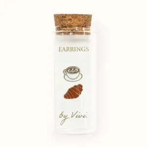 A clear glass phial with a cork lid is labelled Champagne earrings by Vivi. The label is decorated with drawings of a coffee cup, a croissant, and a sparkling champagne flute, hinting at the charming earring designs inside. Gate21.ee