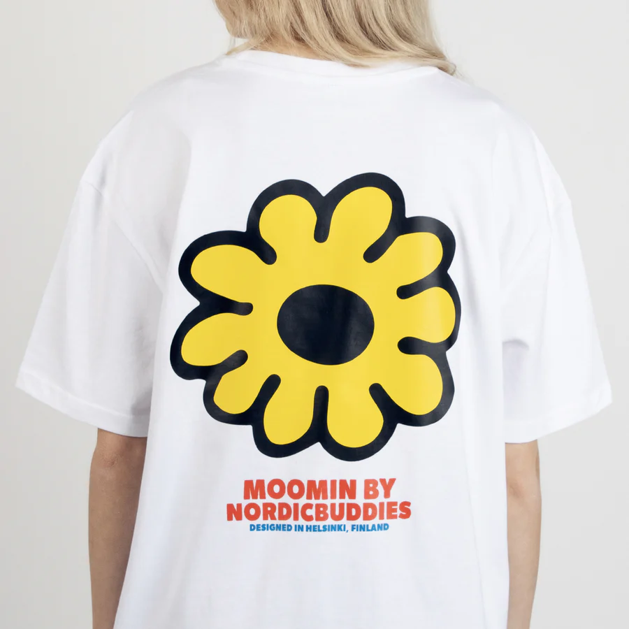 A person wearing the Moomin Ts - Troll’s Flower, a white T-shirt featuring a large yellow flower graphic and “MOOMIN BY NORDICBUDDIES DESIGNED IN HELSINKI, FINLAND” printed in red and blue on the back. Gate21.ee