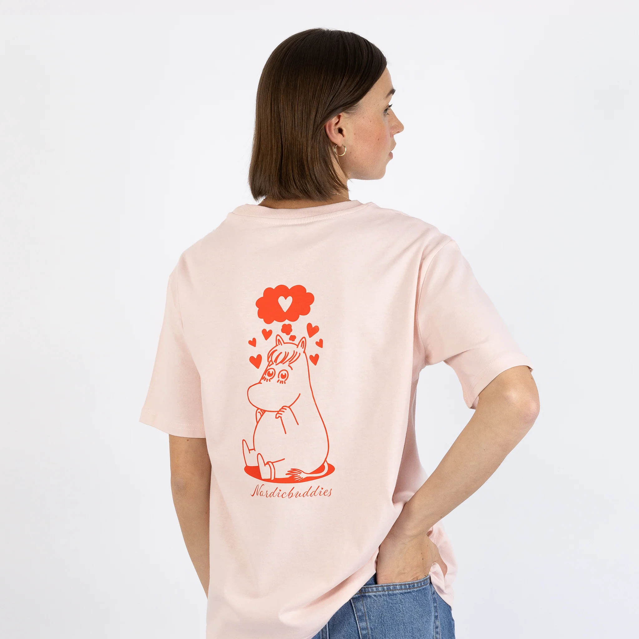 A person stands with their back to the camera, wearing a light pink “Moomin Ts - Snorkmaiden in Love” T-shirt featuring a red illustration of Snorkmaiden with hearts and the word “hobiebuddies” printed below the image. Gate21.ee