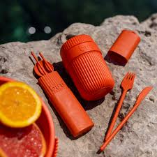 A Bini Cutlery Kit - Sunset (Limited Edition), featuring a set of orange reusable utensils, along with a cup and container, is displayed on a textured rock surface beside slices of citrus fruit in a bowl—creating the perfect limited edition summer essential arrangement. Gate21.ee