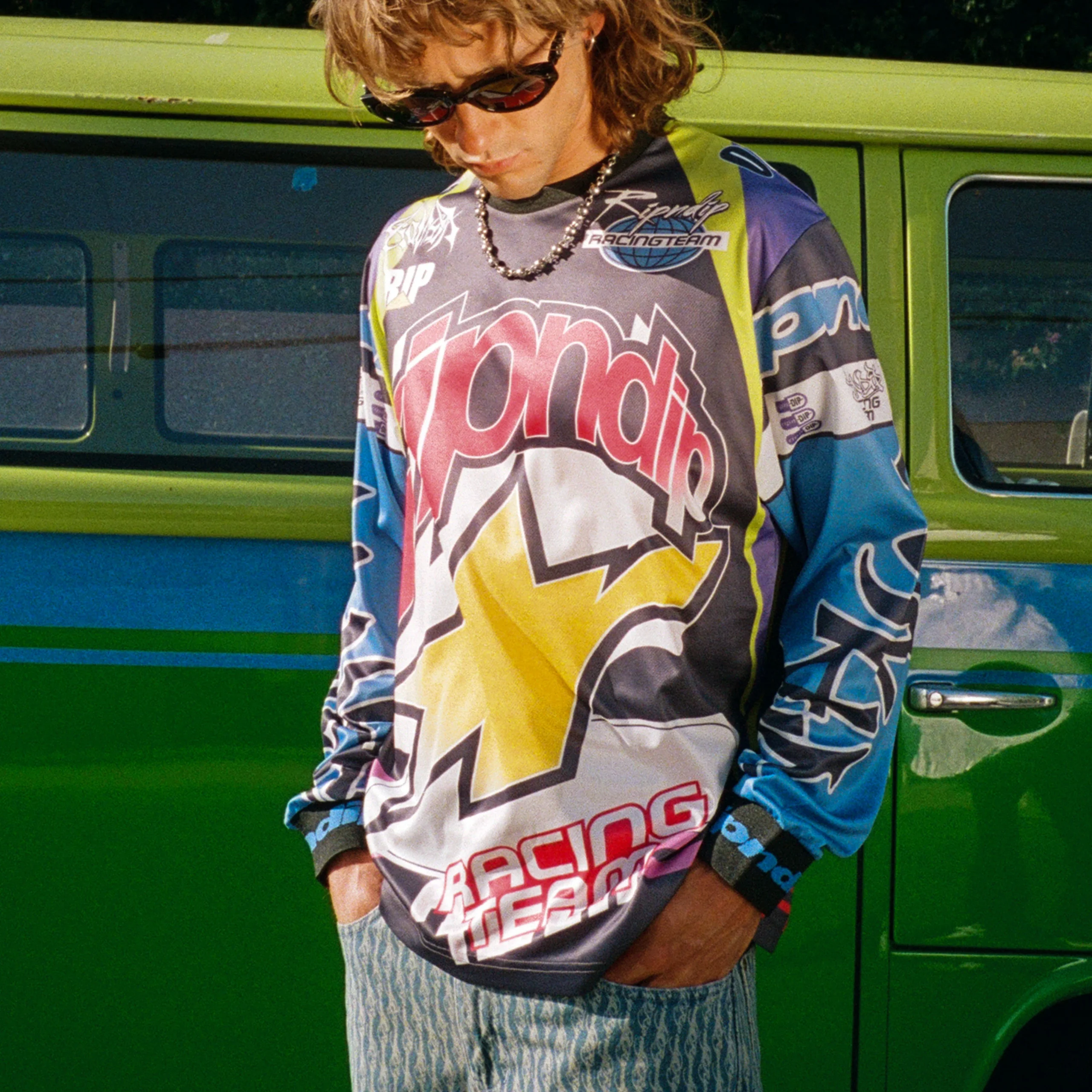 Wearing the Allstar Racing Jersey - Multi-colour from Ripndip, a person with long hair and sunglasses stands in front of a green vehicle, sporting striped trousers and looking down with hands in their pockets. Gate21.ee