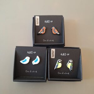 Three pairs of bird-shaped earrings from the ABE jewellery - Birds collection are displayed in elegant black boxes. Each pair features a unique bird design with vibrant colours and distinct styles. Every box is labelled &ldquo;One of a kind&rdquo; and includes a small tag with extra text in the upper left corner. Gate21.ee