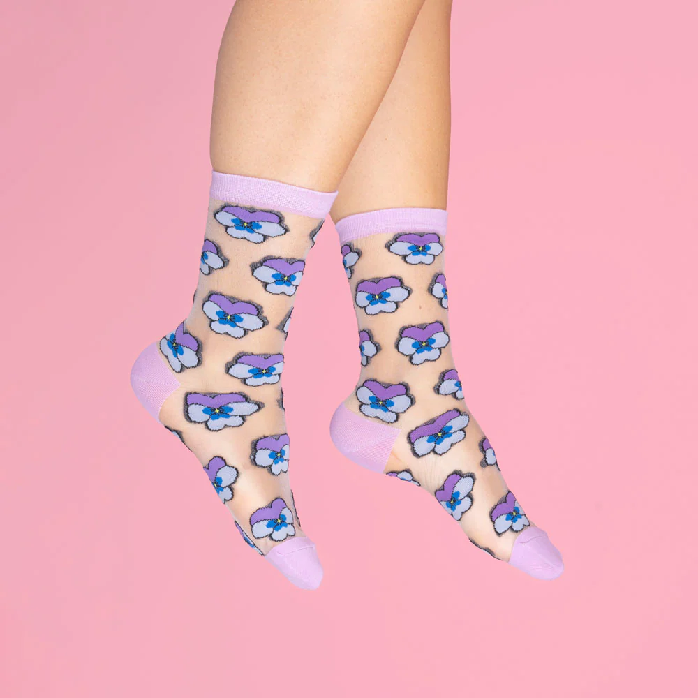 A person wearing the Pansies - Transparent Socks by Coucou Suzette, featuring blue and purple flower patterns with solid lavender toes, heels, and cuffs, is shown against a plain pink background. Gate21.ee