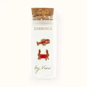 A clear glass vial with a cork lid displays the Champagne earrings by Vivi—two mismatched gold and red earrings in the whimsical shapes of a lobster and a crab. The label reads “Earrings by Vivi,” set against a plain white background. Gate21.ee