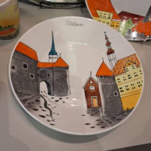 A Helina Tilk bowl features a vibrant illustration of historic buildings with steeples and towers, labelled Tallinn at the top. The design incorporates orange, yellow, grey, and blue tones. Other decorative ceramic pieces are displayed nearby. Gate21.ee